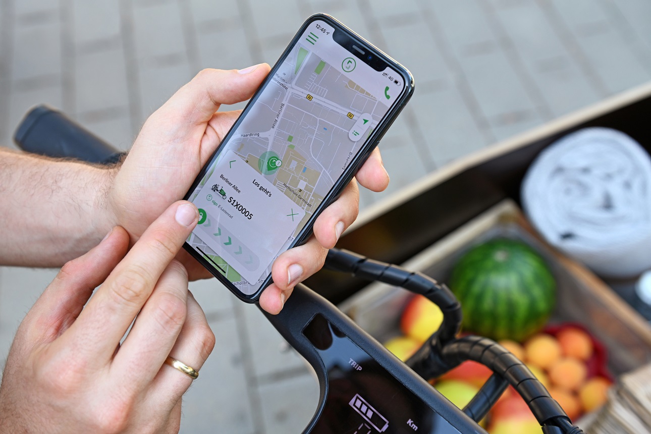 Zu sehen ist eine Hand, die mit dem Handy in der App von Sigo navigiert. Im Hintergrund ist das Lastenrad von sigo angeschnitten.