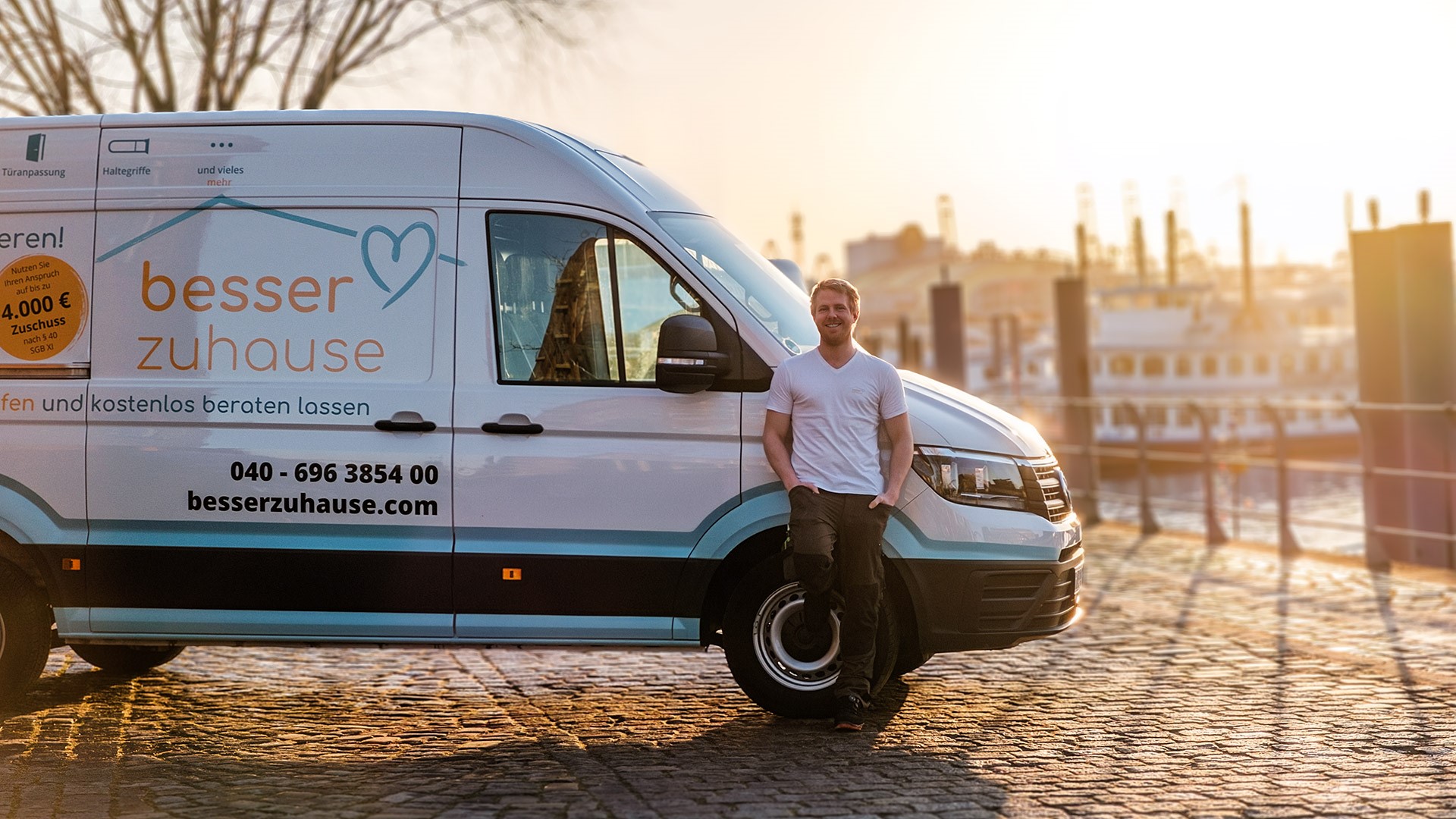 Transporter mit der Werbebeschriftung der besser zuhause GmbH. Davor ein lächelnder Mitarbeiter. Im Hintergrund geht die Sonne unter.