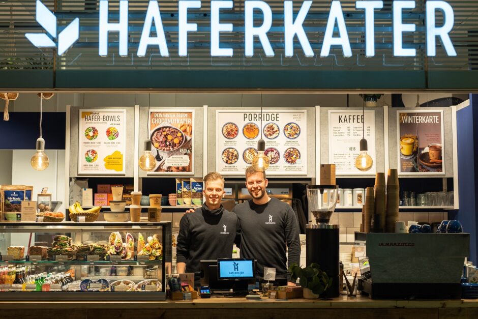 Zwei Mitarbeiter stehen lächelnd hinter der Theke eines Haferkater-Cafés. Das Café bietet Hafer-Bowls, Porridge und Kaffee an, und repräsentiert ein werteorientiertes Geschäftsmodell sowie das Verantwortungseigentum.