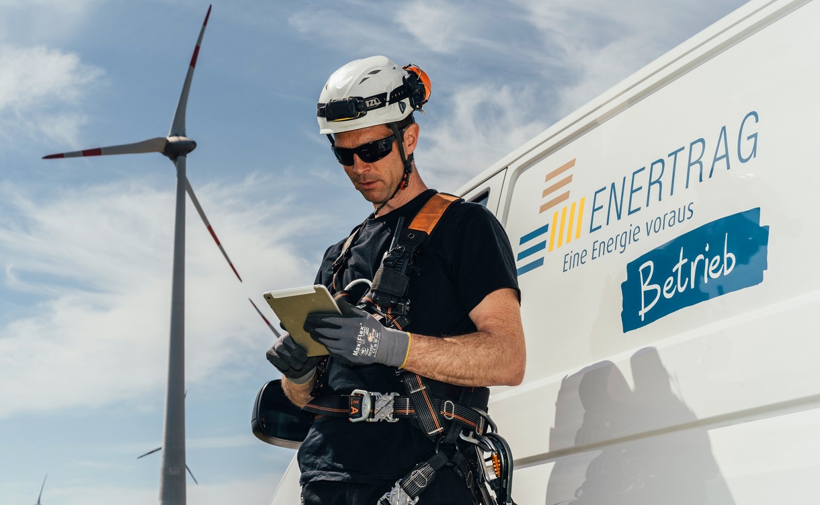 Ein Foto zeigt ein Windrad und einen Monteur des Unternehmens Enertrag, der an einem Tablet arbeitet.
