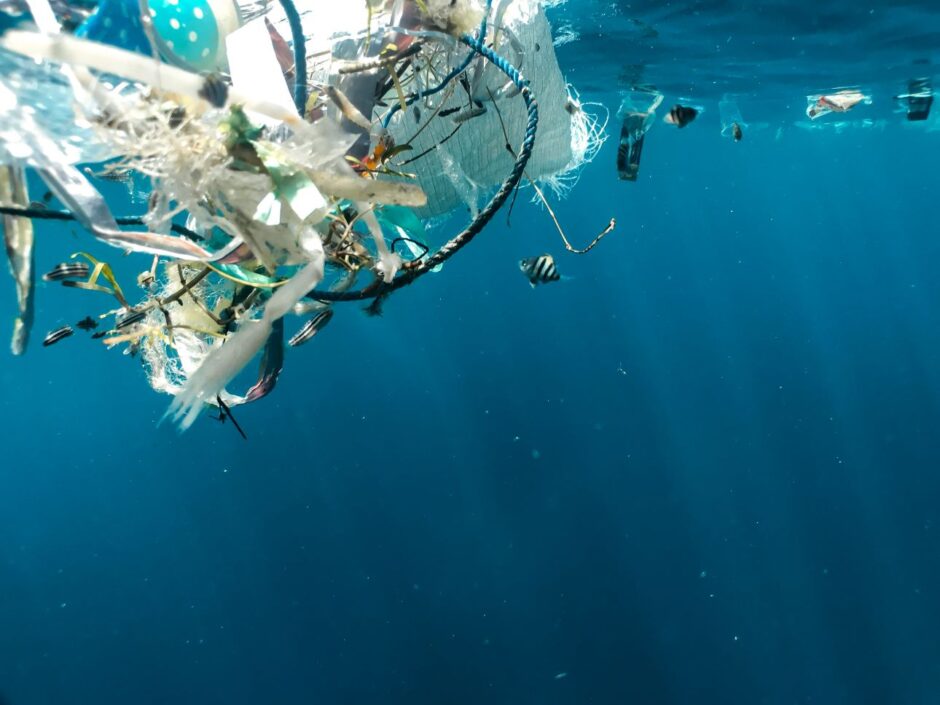 Unterwasseraufnahme von Plastikmüll, der im Meer treibt. Verschiedene Arten von Abfall, darunter Plastikstreifen, Netze und Verpackungen, sind sichtbar. Kleine Fische schwimmen um den Müll herum. Das Bild symbolisiert die Umweltverschmutzung durch Plastik und deren Auswirkungen auf das marine Ökosystem.