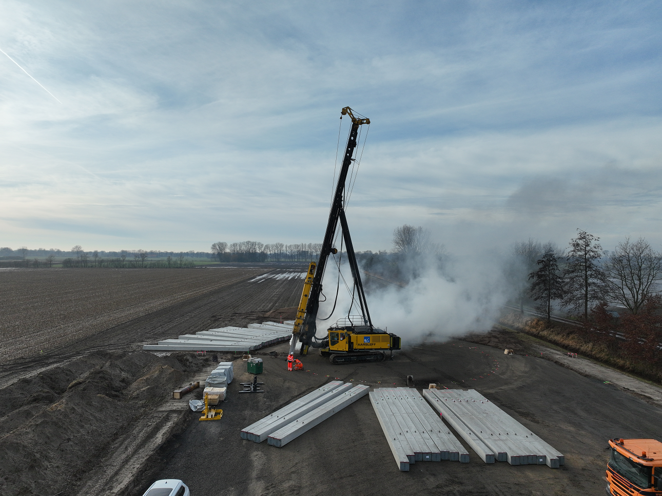 Großes gelbes Drehbohrgerät steht auf dem Baugrundstück auf dem eine Windkraftanlage entstehen soll. Drum herum liegen die Pfähle, die für das Fundament benötigt werden.