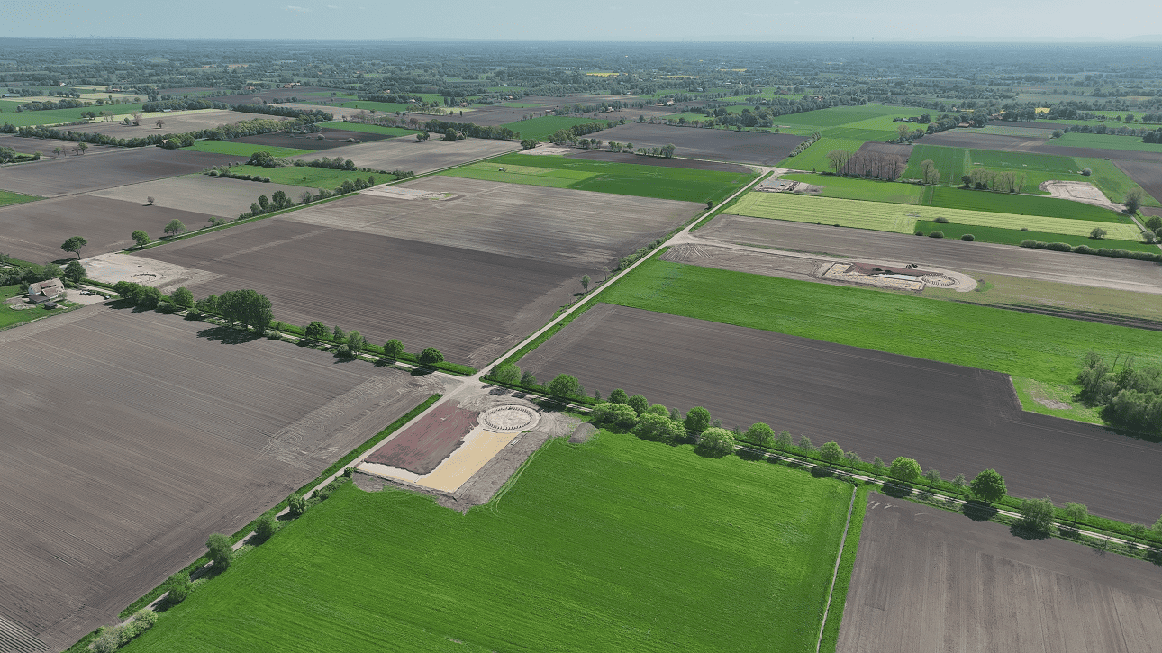 Luftaufnahme einer landwirtschaftlich genutzten Landschaft mit mehreren Feldern und Wegen, im Vordergrund eine Baustelle mit einer kreisförmigen Fundamentstruktur für eine Windenergieanlage.