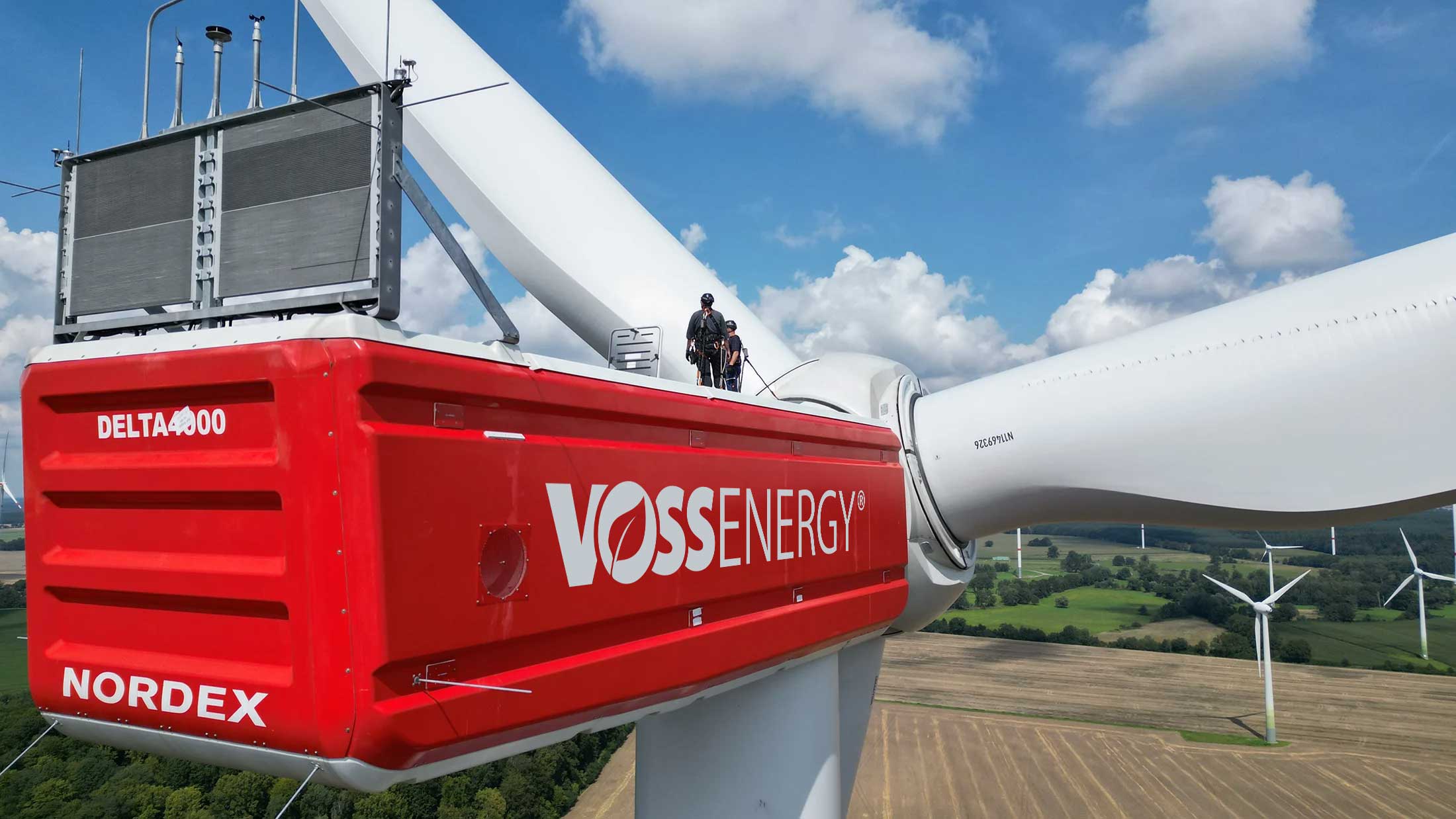 Nahe Luftaufnahme von hinten des Maschinenhauses einer rot lackierten Voss-Windkraftanlage. Auf der Anlage stehen zwei Techniker in Sicherheitskleidung, die Rotornarbe und die Rotorblätter sieht man im Hintergrund.