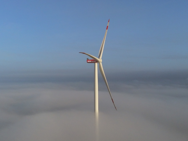 Windkraftanlage der VOSS Beteiligung GmbH ragt aus tief liegenden Wolken heraus, dahinter blauer Himmel.