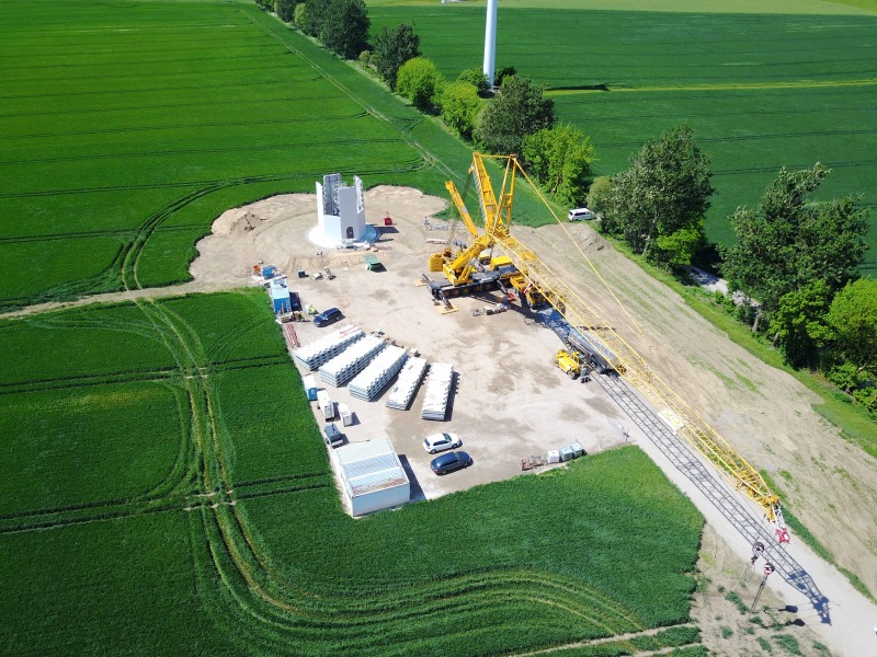 Luftaufnahme der Baustelle eines Windrads. Ein großer gelber Gittermast-Mobilkran hebt die einzelnen Bauteile zur bisher gebauten Anlage. Um die Baustelle herum grüne Felder.