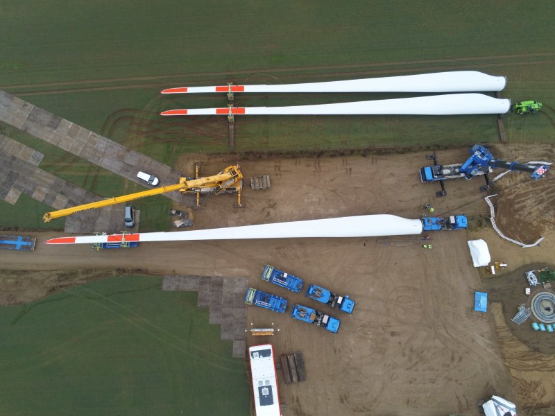 Luftaufnahme von der Anlieferung der Rotorblätter per Lkw für eine Windkraftanlage, sie werden von zwei Kränen abgeladen und am Rand der Baustelle liegend platziert
