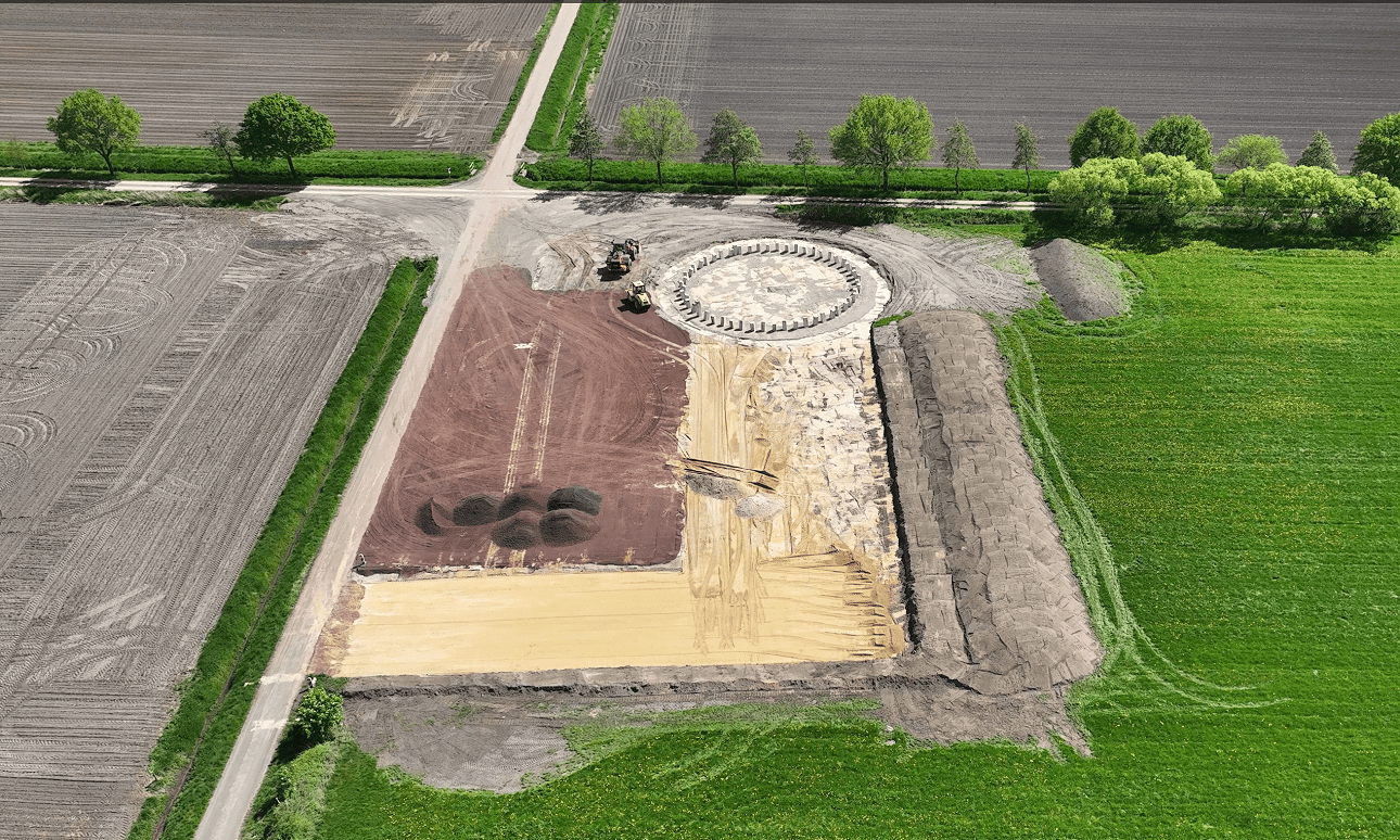 Nahaufnahme einer Baustelle auf einem Feld, auf der ein großes, rundes Fundament für eine Windkraftanlage mit vorbereiteten Betonfundamenten und Baumaschinen zu sehen ist.