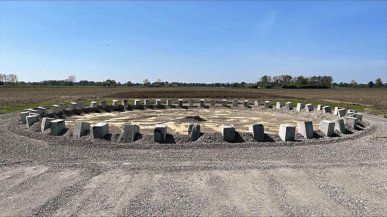 Bodensicht auf das runde Fundament einer Windkraftanlage, bestehend aus mehreren aufgestellten Betonblöcken im Kreis auf einer Baustelle inmitten von Feldern.