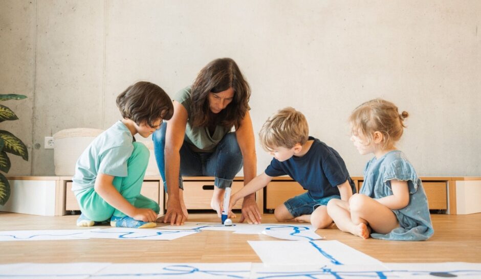 Eine erwachsene Person sitzt mit drei kleinen Kindern auf einem Holzboden in einem hellen Raum einer Kita von littlebigFuture. Die Gruppe beugt sich vor und beschäftigt sich gemeinsam mit einem kreativen Malprojekt auf großen weißen Papierbögen, auf denen blau gemalt wurde. Die Kinder tragen bequeme Freizeitkleidung und sind aufmerksam bei der Aktivität. Im Hintergrund steht ein Sideboard mit einigen Pflanzen am Rand. Die Szene wirkt freundlich und konzentriert, im Stil einer spielerischen Lernumgebung.